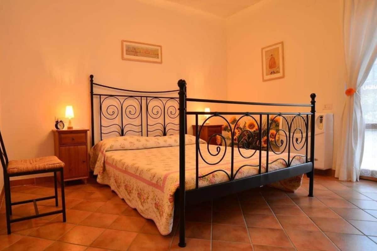 Een slaapkamer met een metalen bedframe, een nachtkastje met een lamp, een stoel en een raam met gordijnen.
