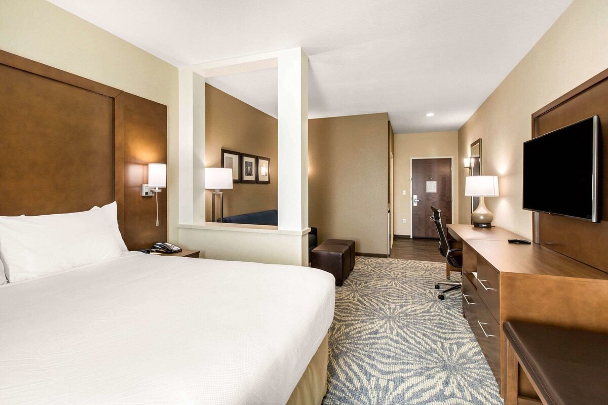 A hotel room with a large bed, a desk with a flat-screen TV, a mirror, and a door leading to another room.