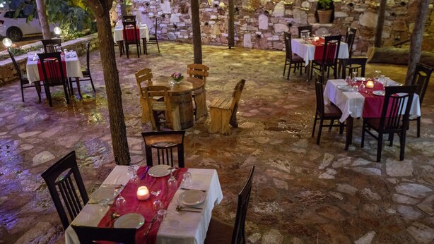 An outdoor dining area with stone flooring and tables set for two, surrounded by stone walls and trees. - Hotel Horizont (Aliko)