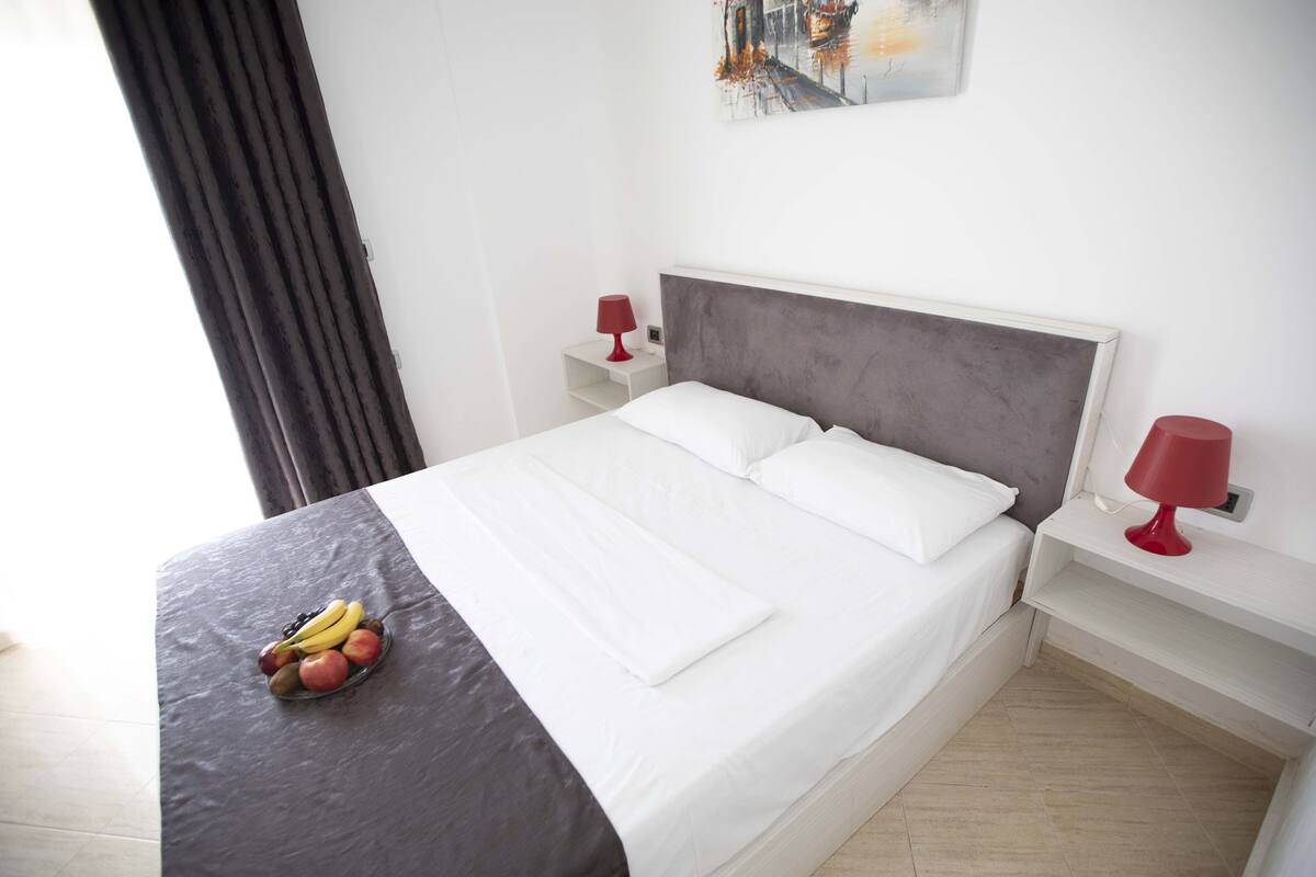 A neatly made bed with white linens, two bedside tables with red lamps, a fruit bowl on the bed, and a painting on the wall.