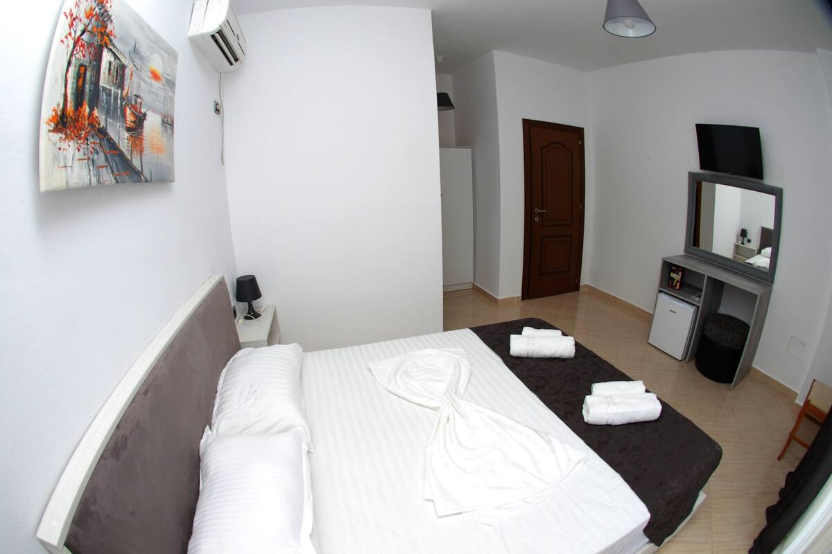 A hotel room with a bed, a desk, a mirror, and a small refrigerator.