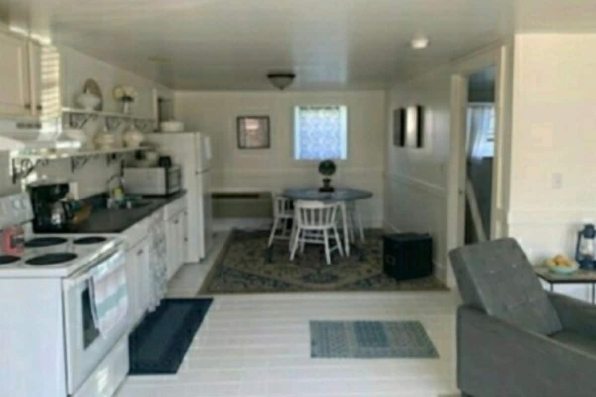 A compact kitchen with white appliances, a dining area with a small table and two chairs, and a grey sofa in a living space.