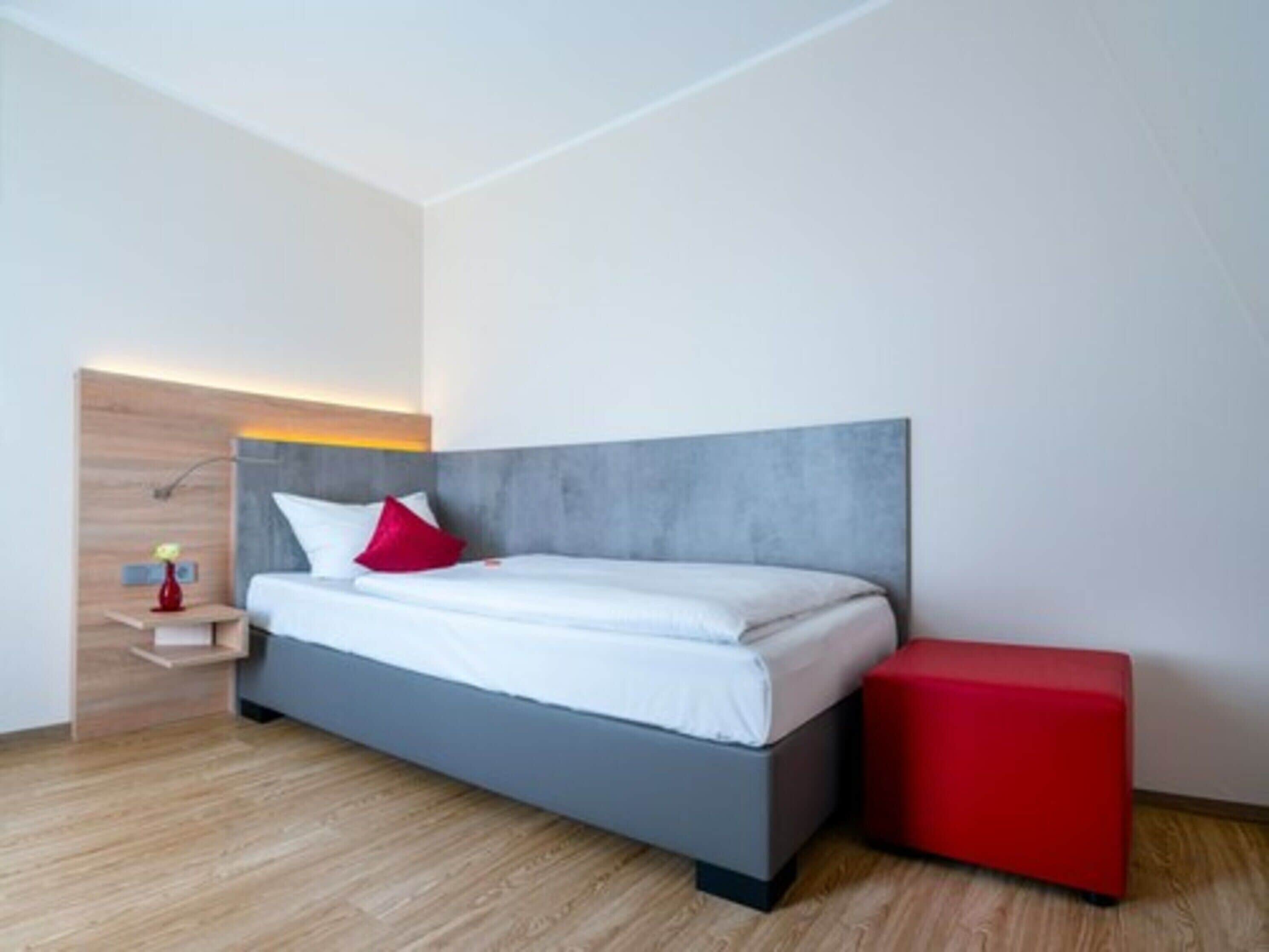 A modern bedroom with a grey bed, white bedding, a red pillow, a bedside table with a lamp, and a red ottoman.