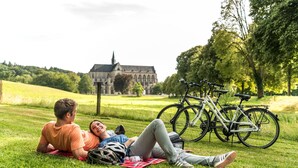 Two people lying on a blanket in a grassy area with bicycles and a historic building in the background. - Landhotel Fettehenne (Leverkusen)