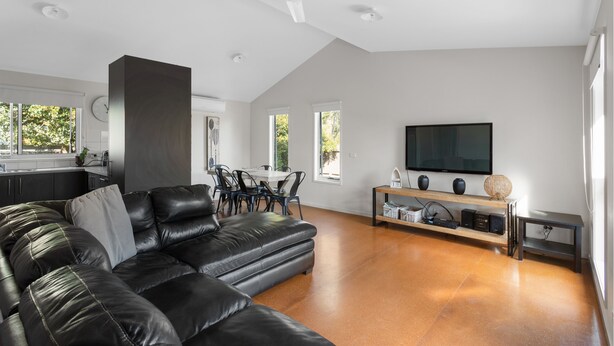 A modern living room with a black leather sofa, a dining area with a white table, and a flat-screen TV mounted on the wall. - Cowes Retreat - *Pet Friendly* Holiday House!!! (Cowes)