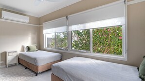 A bedroom with two beds, a ceiling fan, an air conditioner, and large windows with a view of trees and flowers.