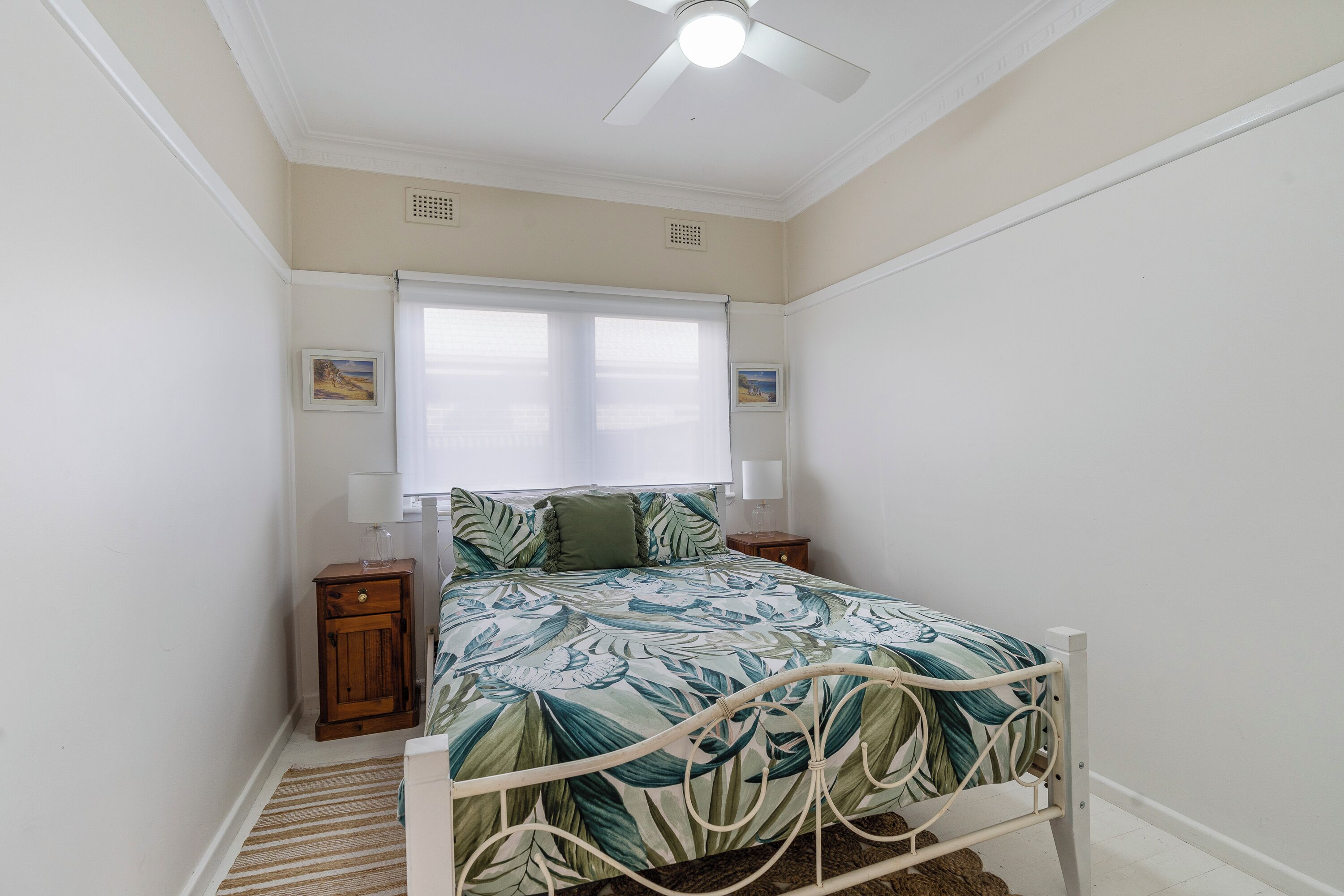 Un dormitorio con cama, ventilador de techo, ventana con persianas y mesita de noche.