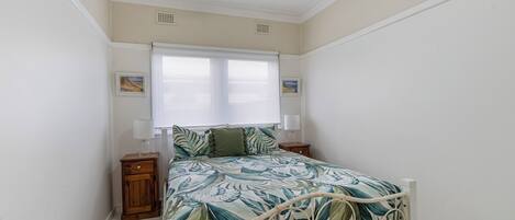 Un dormitorio con cama, ventilador de techo, ventana con persianas y mesita de noche.