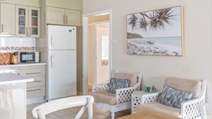 A kitchen with white cabinets, a microwave, and a refrigerator. There is a dining table with chairs and a painting of palm trees on the wall.