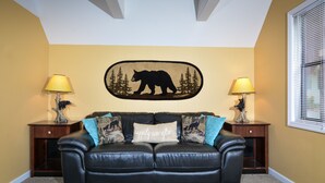 A black leather sofa with a bear-themed pillow, two wooden nightstands with lamps, a bear silhouette wall art, and a window with blinds. - Gatlinburg Cabin Free Tickets, Heart Whirlpool, Hot Tub, King Bed, Fast Internet (Gatlinburg)
