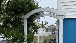 A pathway leading to a patio area with a white picket fence, a blue door, and a view of a residential street. - Lower deck, Salisbury Beach, Melo's Beach House Rentals  (Salisbury)