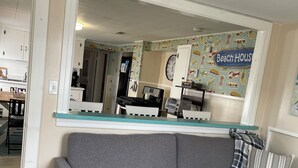 A living room with a grey sofa, a glass-top coffee table, a patterned rug, and a kitchen area visible through a large mirror. - Lower deck, Salisbury Beach, Melo's Beach House Rentals  (Salisbury)