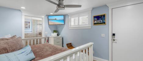 Une chambre à coucher comprenant un lit, un ventilateur de plafond, une télévision, une fenêtre avec des stores et une porte.
