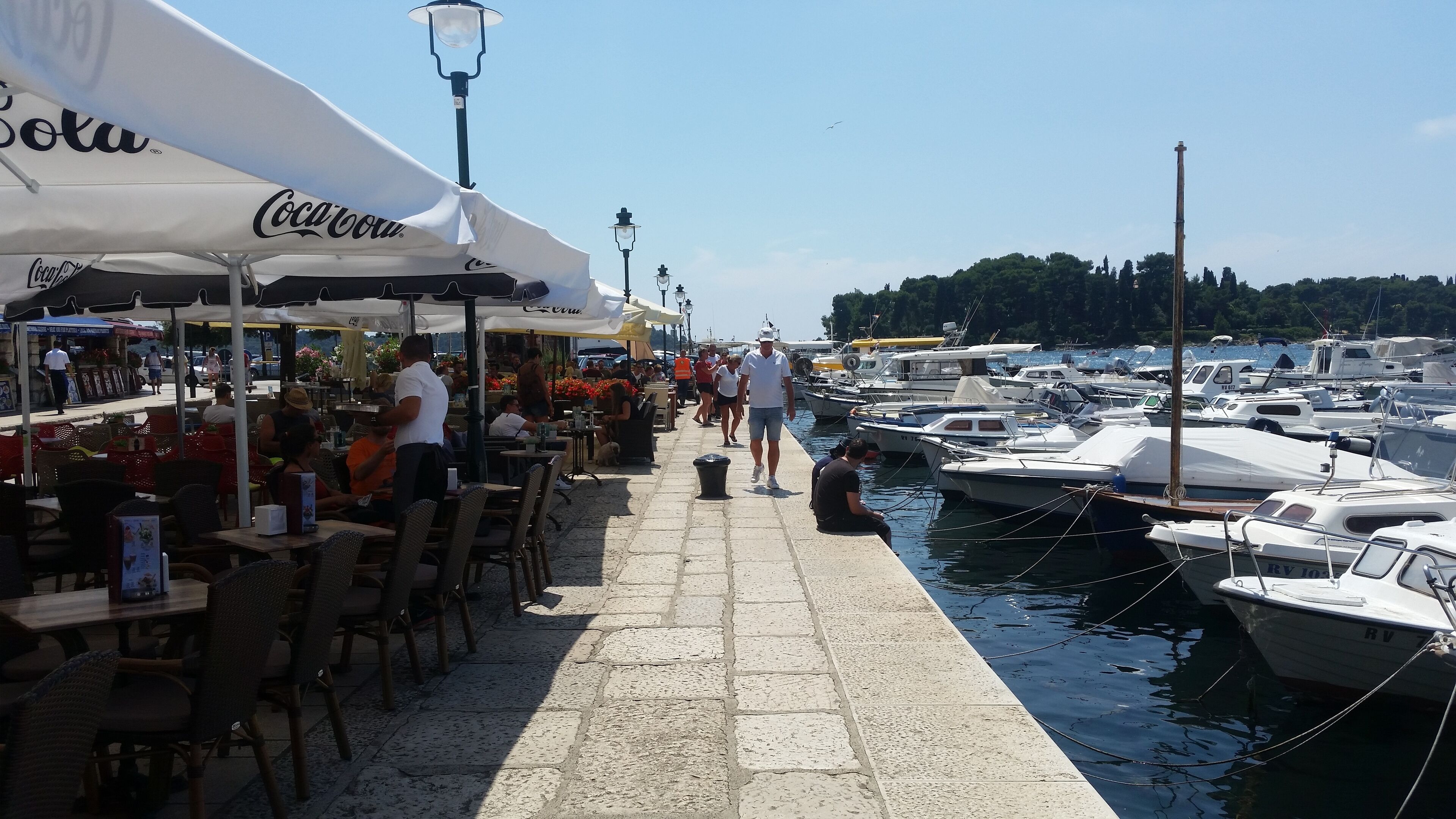 Un port de plaisance avec des bateaux amarrés, une promenade dallée et des espaces de détente en plein air avec des parasols.