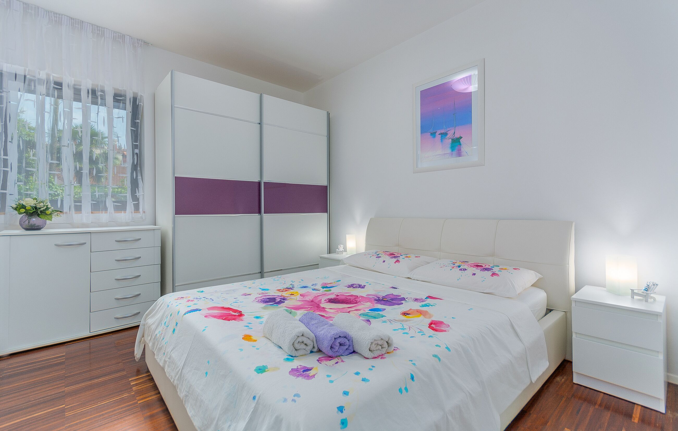 Un dormitorio con cama, armario, mesita de noche y ventana con cortinas.