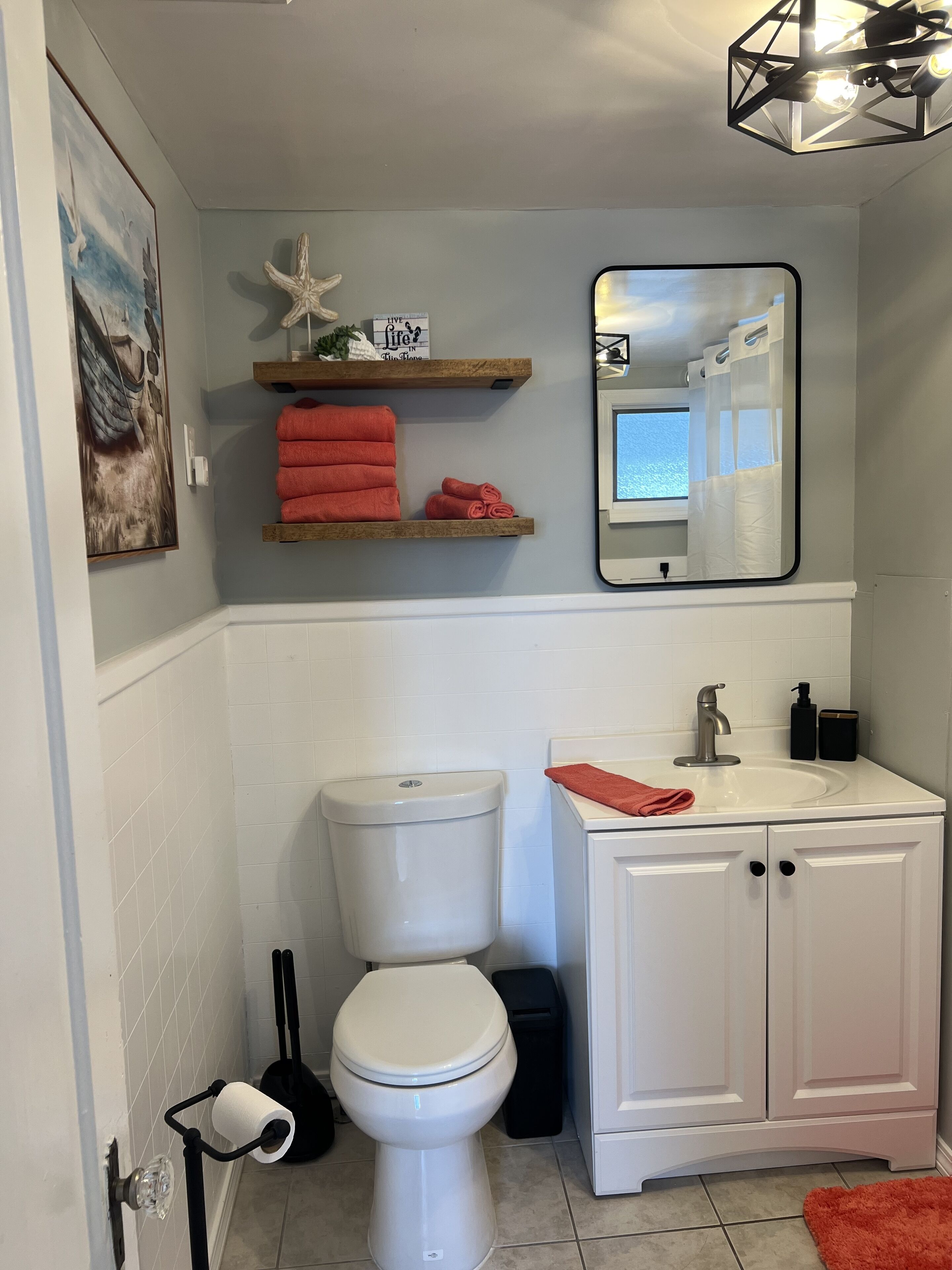 Et badeværelse med toilet, vask og spejl. Der er håndklæder på en hylde, og et billede på væggen.