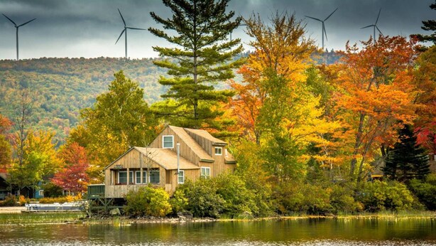 A wooden cabin by a lake with a view of wind turbines in the background.