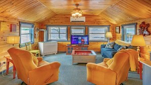 A cozy cabin interior with wooden walls, a central seating area with orange chairs, a television, and a window with a view of the outdoors.