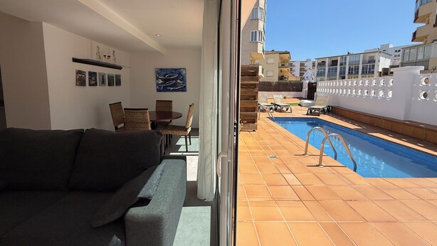 Ein Wohnzimmer mit einer Couch, Esstisch und Stühlen, ein Pool mit Liegestühlen und Blick auf Gebäude.