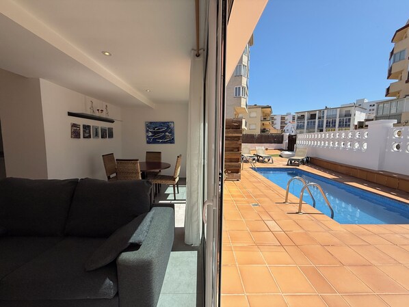 A living room with a sofa, dining table, and chairs, a pool with lounge chairs, and a view of buildings.