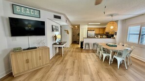 A modern kitchen with wooden cabinets, a dining area with white chairs, and a large flat-screen TV mounted on the wall. - FREE FUN during your stay in this Gold-rated condo with beautiful ocean view and easy access to the pool and beach! (Saint Augustine)