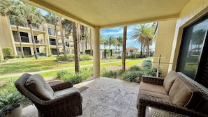 A covered outdoor seating area with wicker furniture, a view of a building with balconies and palm trees, and a pool in the background. - FREE FUN during your stay in this Gold-rated condo with beautiful ocean view and easy access to the pool and beach! (Saint Augustine)