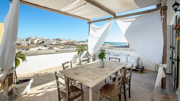 A rooftop terrace with a wooden table and chairs, a potted plant, and a view of the cityscape and sea. - Ortigia Boutique Terrace Apt (Siracusa)