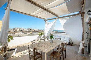 A rooftop terrace with a wooden table and chairs, a potted plant, and a view of the cityscape and sea.