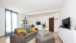 A modern living room with a grey sofa, a laptop on a grey ottoman, a TV on a white cabinet, and a door with a coat rack. - Ortigia Boutique Terrace Apt (Siracusa)