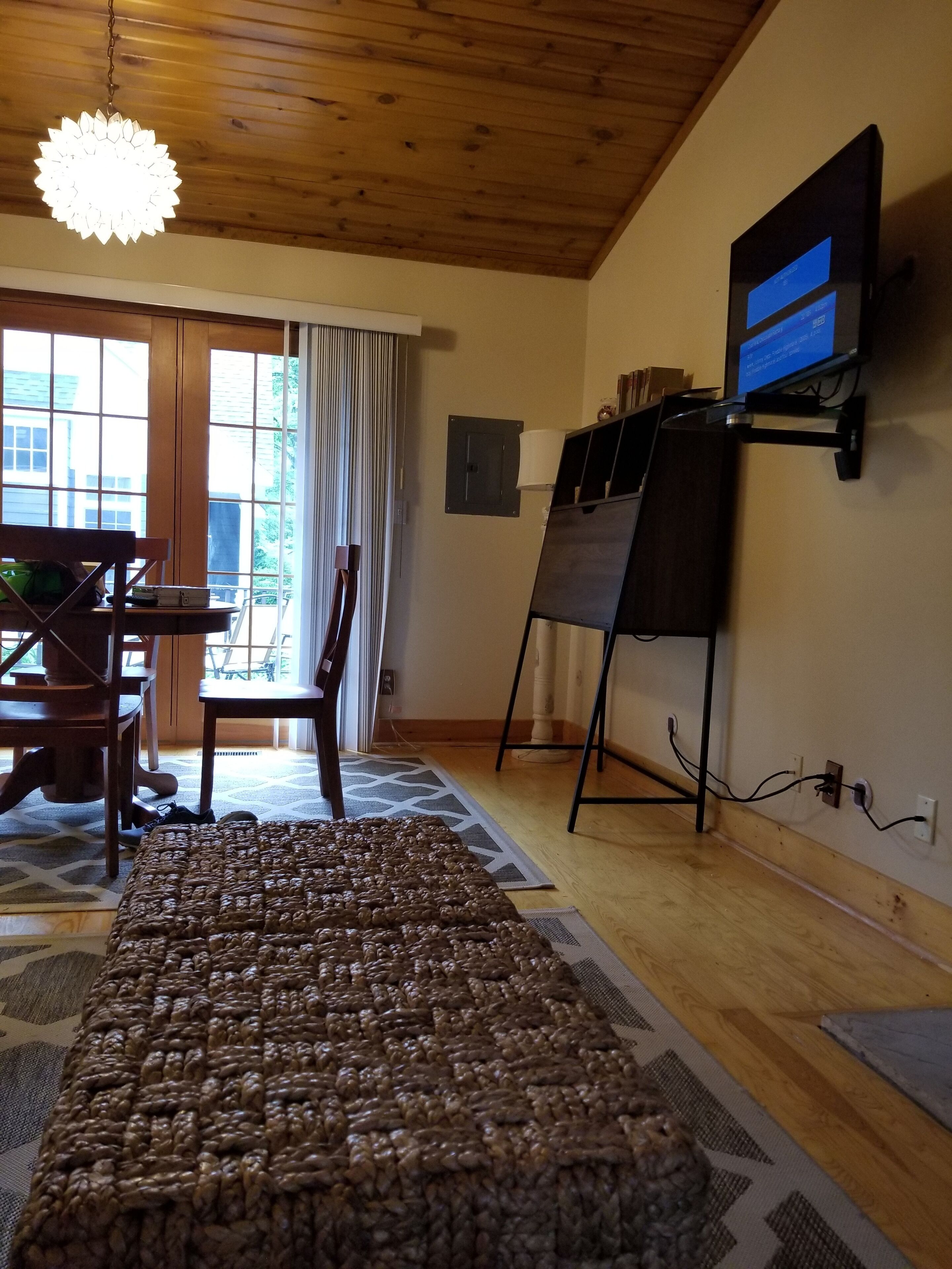 Ein Zimmer mit Holzboden, einem großen Fenster mit Vorhängen, einem Esstisch mit Stühlen, einem Teppich und einem an der Wand befestigten Fernseher.