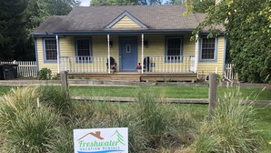 A yellow house with a blue door and a porch. There is a sign in front with the text "Freshwater Vacation Rentals" and a phone number. - Harbor Haven: Lake Michigan Beach Cottage | Union Pier | Family-Friendly (Union Pier)