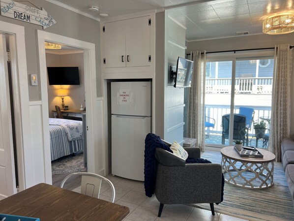 A compact hotel room with a kitchenette, a dining area, a living area with a view of the balcony, and a television mounted on the wall.