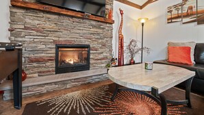 Un salón con chimenea de piedra, un televisor de pantalla plana montado encima, una mesa redonda y un sofá con un cojín rojo.