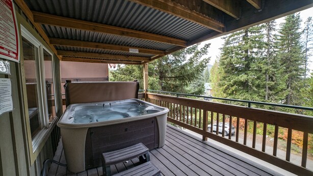 Un bain à remous sur une terrasse en bois, avec vue sur les arbres et une voiture au loin.