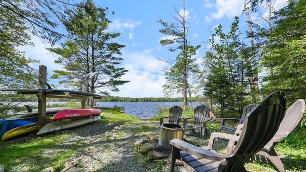 A lakeside seating area with Adirondack chairs and a fire pit, surrounded by trees and a dock with canoes. - DOCKSIDE DAYS-LAKEFRONT in Lake Naomi!  Boating, Sunsets and Year-Round Fun (Pocono Pines)