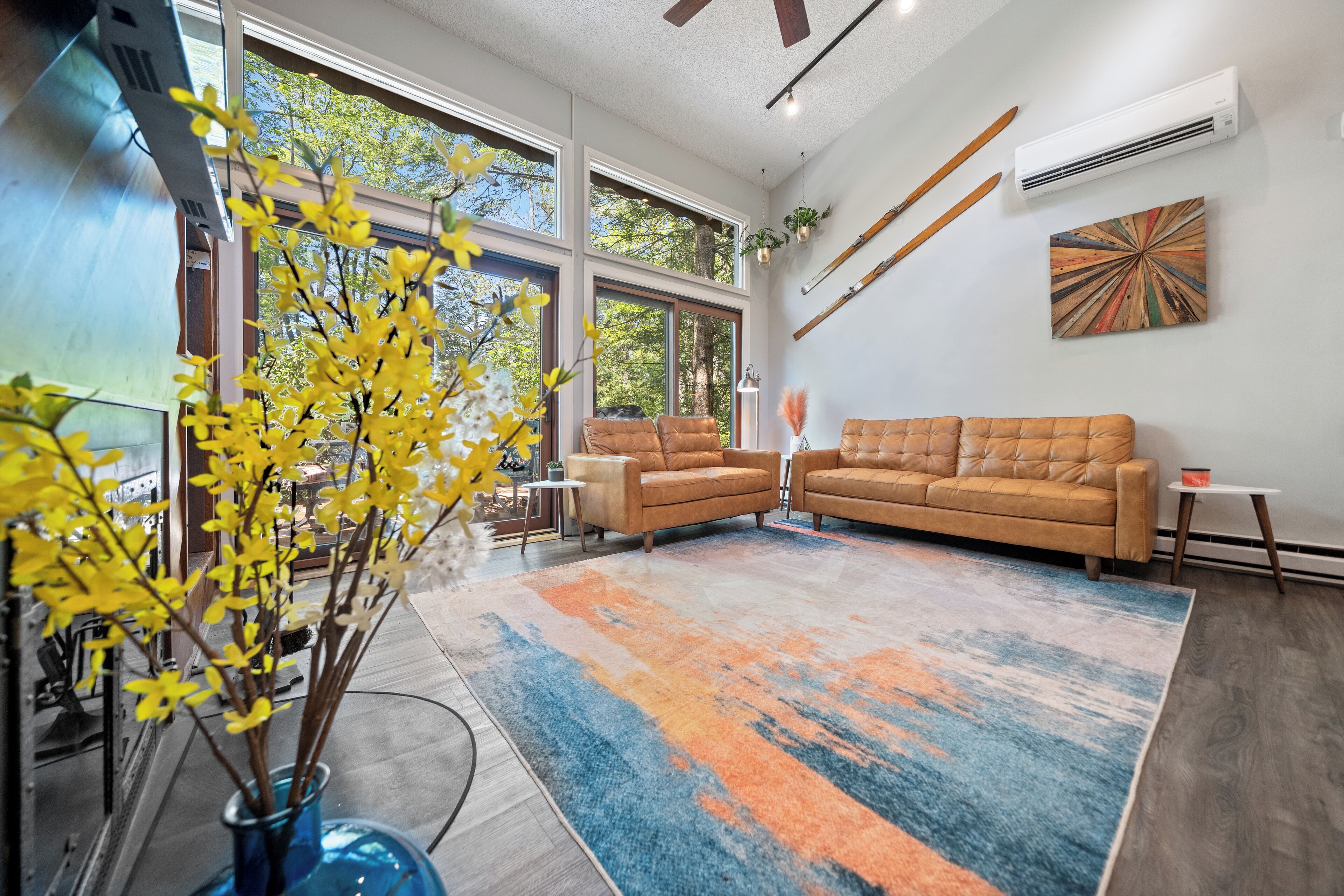 Un salon moderne avec de grandes fenêtres, un canapé en cuir marron, un tapis coloré et un vase bleu contenant des fleurs jaunes.