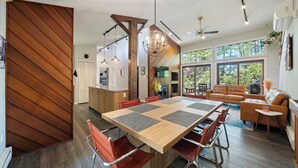 A spacious living area with a large wooden dining table, modern furniture, and large windows offering a view of the outdoors. - DOCKSIDE DAYS-LAKEFRONT in Lake Naomi!  Boating, Sunsets and Year-Round Fun (Pocono Pines)