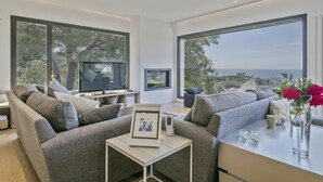 Ein modernes Wohnzimmer mit einer Couch, einem Couchtisch und einem Fernseher. Große Fenster bieten Blick ins Freie.
