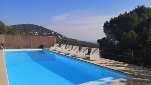 Ein rechteckiger Swimmingpool mit klar blauem Wasser, umgeben von einer Steindeck. Entlang des Poolrandes befinden sich mehrere Liegestühle, und ein Holzzaun trennt den Poolbereich von einem Hügel mit Gebäuden im Hintergrund.
