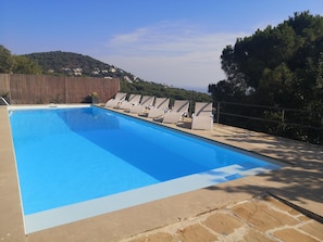 A rectangular swimming pool with a clear blue water surface, surrounded by a stone deck. There are several lounge chairs aligned along the pool's edge, and a wooden fence separates the pool area from a hillside with buildings in the background. - DESMAIS design house and breathtaking views (Cabrils)