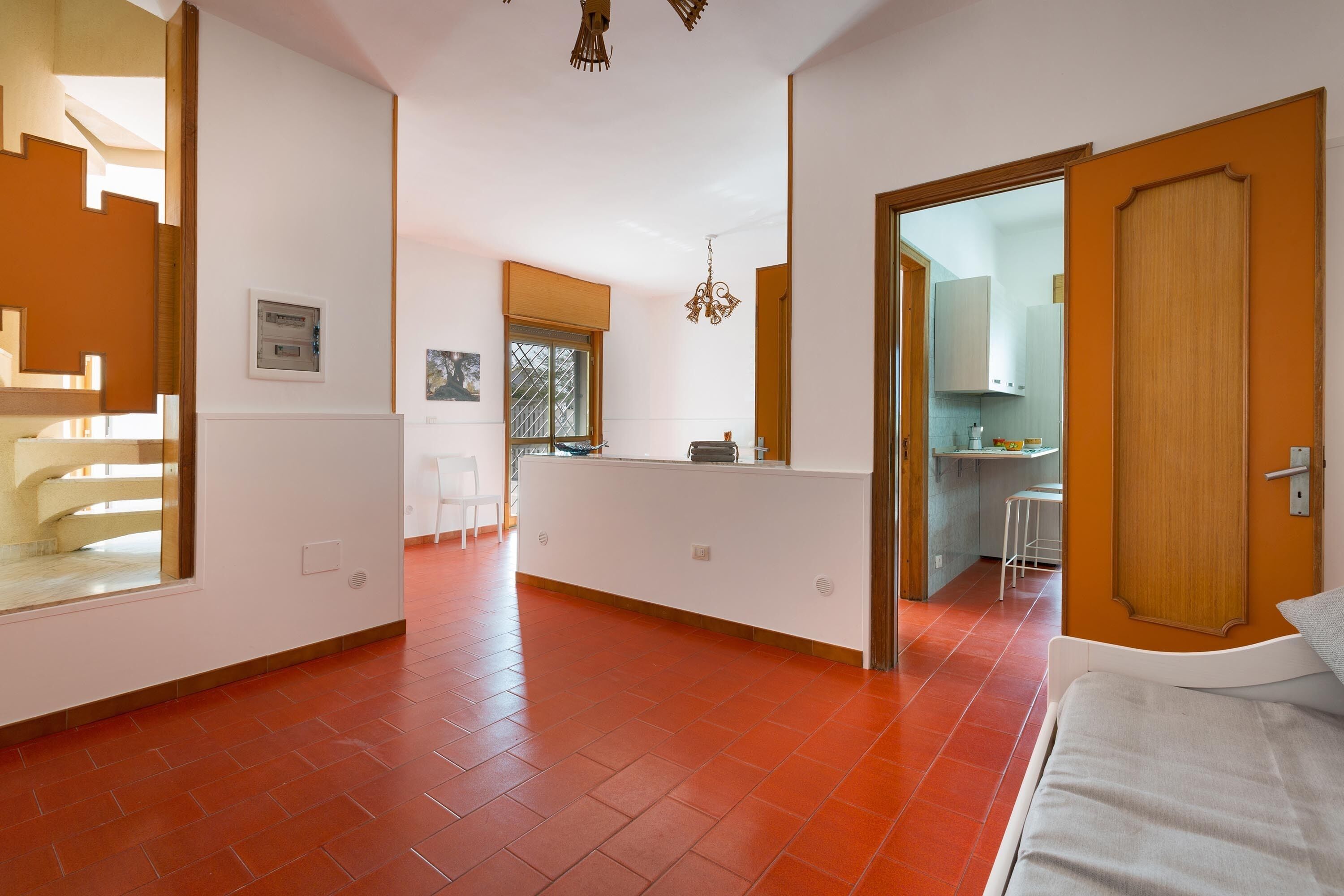 Habitación de hotel moderna con piso de baldosas rojas, una cama blanca y vista hacia la zona de la cocina.