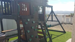 Een houten speelconstructie met klimelementen en een glijbaan, gelegen op kunstgras met uitzicht op verre bergen.
