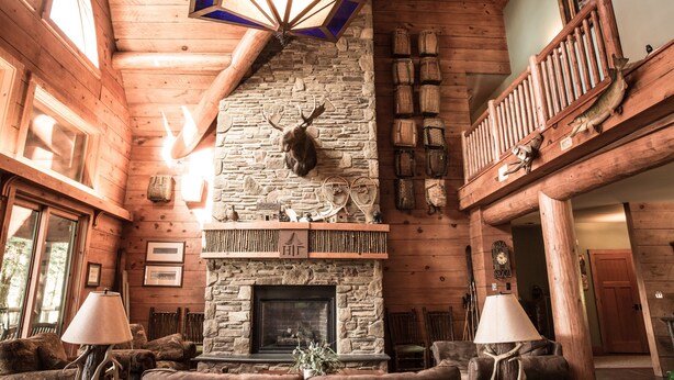 A log cabin interior with a stone fireplace, wooden walls, and a balcony with fishing lures.