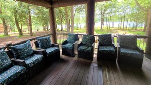 A covered outdoor seating area with wicker furniture and patterned cushions, overlooking a lake and forested area.