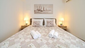 Una cama bien hecha con colcha floral, dos almohadas con diseños estampados y toallas enrolladas sobre la cama. Hay dos lámparas de noche a cada lado de la cama, y un cuadro con barcos de vela en la pared de arriba.