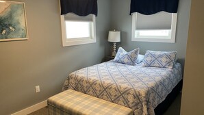Une chambre à coucher avec un lit, deux fenêtres équipées de rideaux foncés, une table de chevet avec une lampe et un tableau accroché au mur.