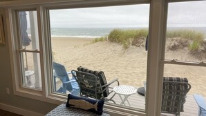 Ett rum med stora fönster med utsikt över en strand, inrett med en blå fåtölj, en mönstrad pall och ett litet runt bord.