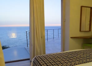 A room with a large window overlooking the sea, a bed with a patterned bedspread, and a wooden shelf.