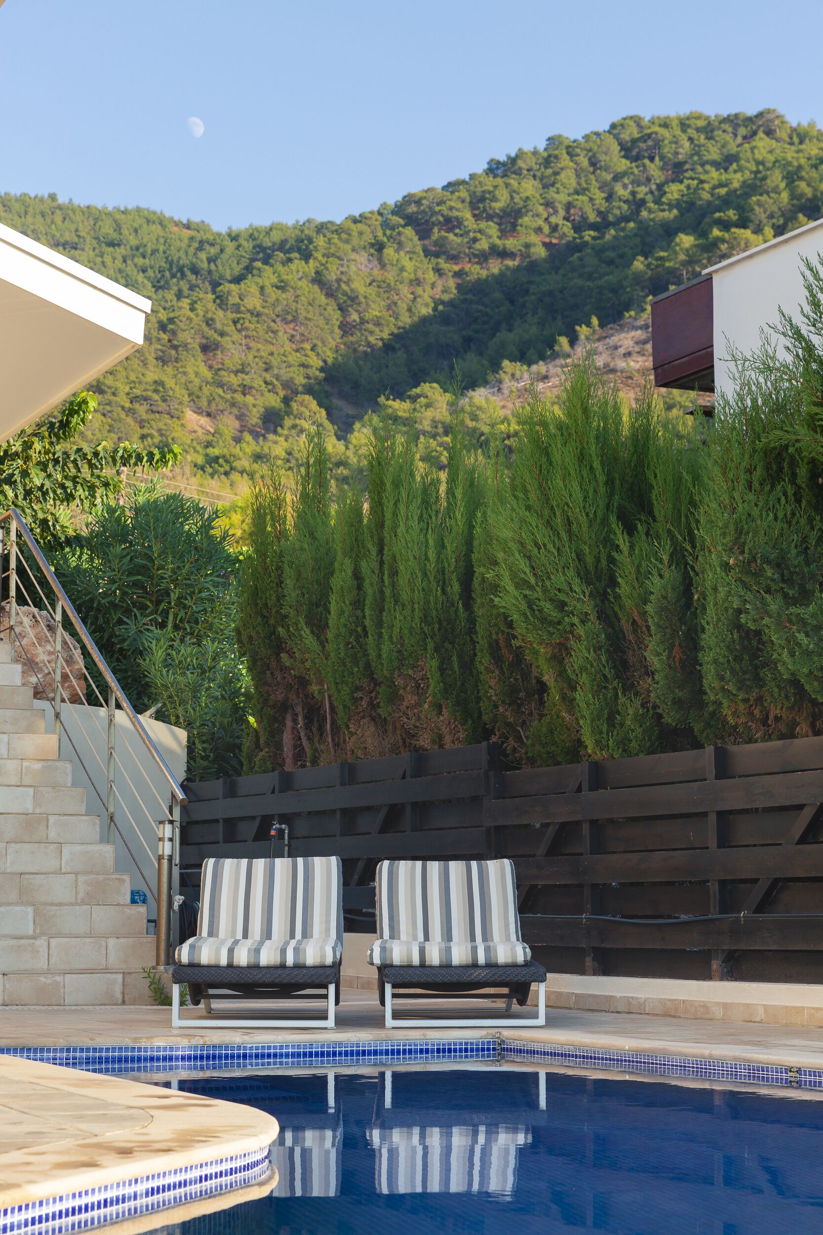 Zona de piscina con dos sillas de descanso, una terraza de madera y vistas a un paisaje montañoso.