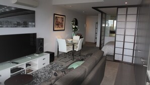 A modern living room with a flat-screen TV, a sofa, a dining area with a table and chairs, and a sliding glass door leading to a bedroom. - Luxurious with great city views, free Private parking & fast internet  (South Yarra)